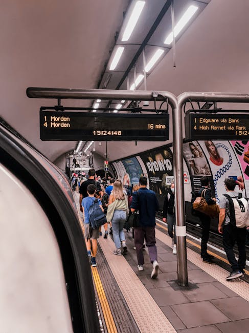 A close-up view of a London Underground sign mounted on a pole in front of a building with reflective glass windows. The sign features a red circular frame with a blue bar across the middle displaying