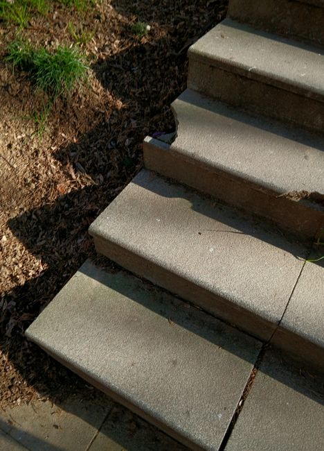 Close-up view of three concrete steps leading up to an entrance, with the bottom step casting a shadow on the ground. To the left side, there is a patch of soil with some green grass and small plants. Inside the property, a wooden floor and cardboard moving boxes are visible, indicating a home relocation process. The boxes are stacked near the steps, and a person is seen lifting or carrying a large wrapped item, likely furniture, wearing dark clothing. The scene appears to be outdoors, on a paved pavement or driveway adjacent to the entrance. The environment suggests ongoing internal and external furniture transport as part of a moving service provided by Man with Van Morden Park, focused on navigating tight staircases in Morden Park flats in the process of packing, loading, and moving household items.
