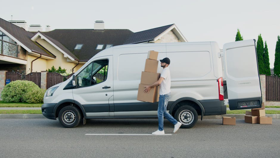 A person wearing a red vest, black trousers, and a blue beanie is seen unloading cardboard boxes from the back of a small light grey panel van parked in an empty parking lot outside a modern, white co