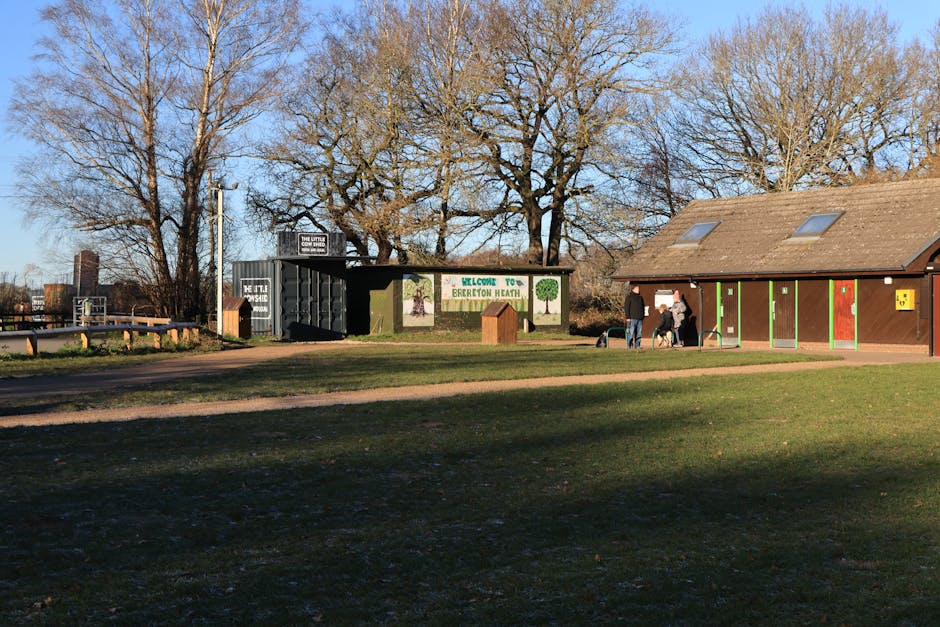 A wide asphalt pathway lined with large deciduous trees, some with yellowing leaves, extends through a park-like setting on a clear day. Wooden benches with metal armrests and legs are positioned along the grassy edges of the path, casting shadows on the ground. In the background, residential houses with brick facades and pitched roofs are visible behind the trees. The scene is well-lit, with sunlight filtering through the tree branches, creating dappled patterns of light and shadow on the pathway. This peaceful outdoor environment may serve as an area where property owners, supported by companies like Man with Van Morden Park, coordinate home relocation or furniture transport activities outdoors, during which they might load or unload household items such as boxes and furnishings from moving vehicles, although no people or moving equipment are visible in this image.