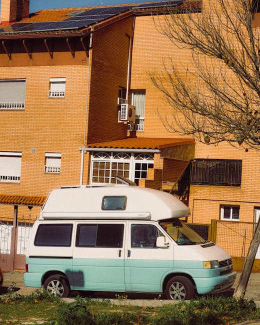 A white and light turquoise van with a high roof is parked on the street in front of a multi-storey brick residential building, part of a home relocation process in progress. The van's side windows an