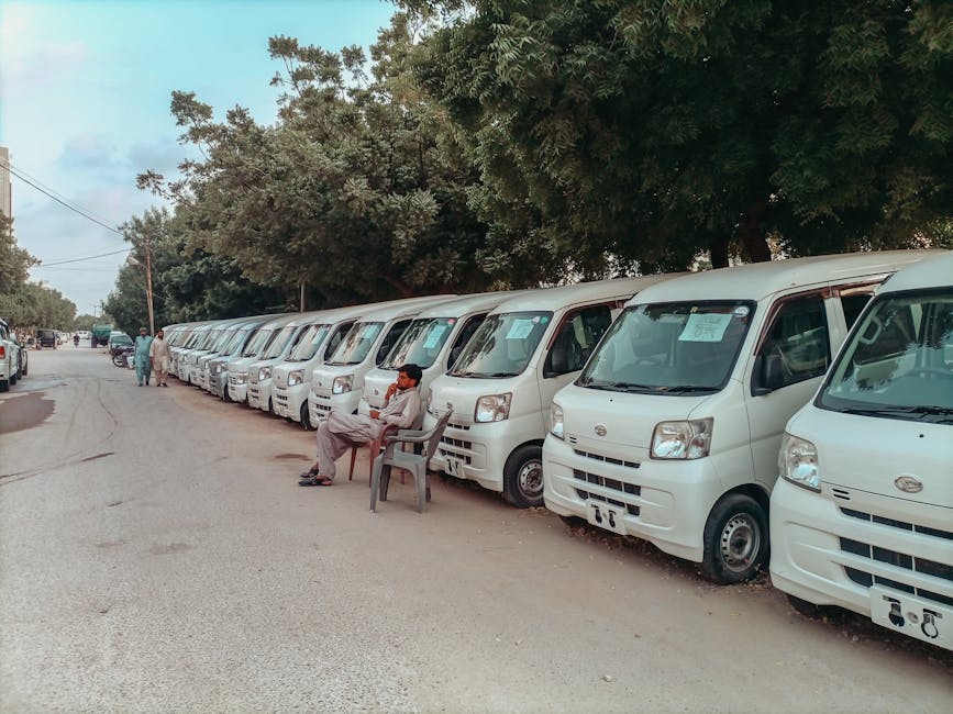 A white and light turquoise van with a high roof is parked on the street in front of a multi-storey brick residential building, part of a home relocation process in progress. The van's side windows an