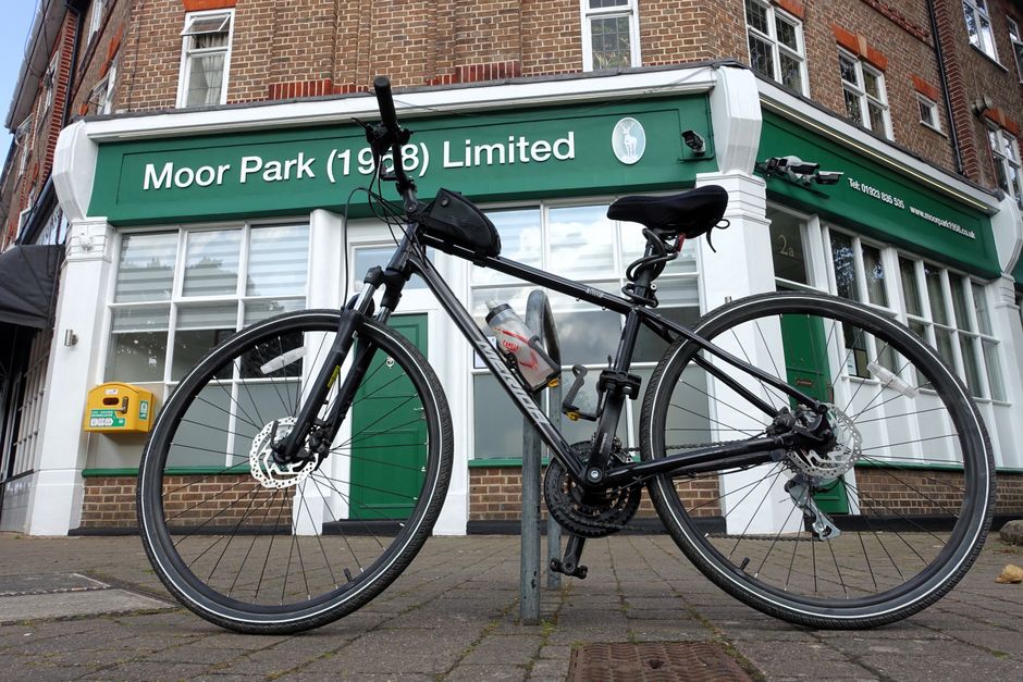 A black mountain bike with a water bottle mounted on the frame is secured to a metal pole in front of a brick building housing Moor Park (1980) Limited, a bakery and café. The bike features front sus