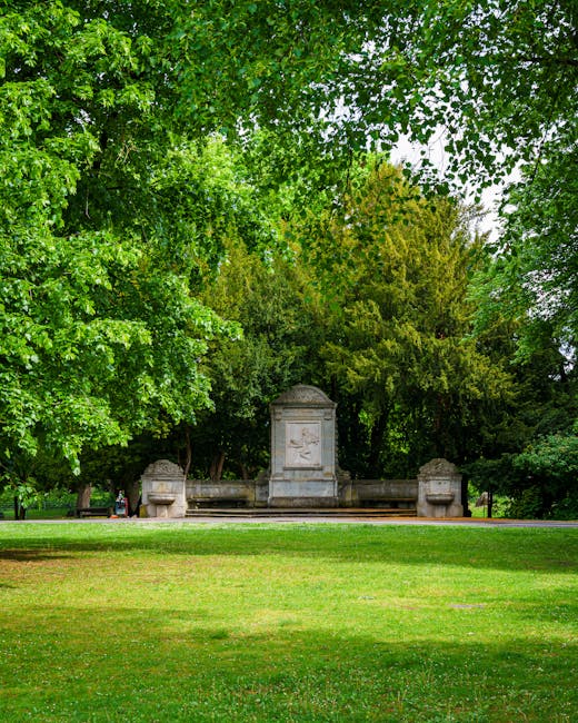 A peaceful park scene with a green lawn in the foreground and tall trees with lush foliage forming a canopy overhead. In the middle distance, there is a historic stone monument with intricate carvings, flanked by two stone benches. The monument and benches are situated on a paved pathway, which appears to be part of a larger park or public garden. The area is well-maintained and shaded by the surrounding trees, providing a tranquil environment suitable for relaxing or outdoor activities. This setting exemplifies a typical outdoor location for house removals and relocation services, where items might be temporarily stored or loaded onto vehicles during a furniture transport or home moving process. The natural light highlights the greenery and stone features, emphasizing the calm and organized atmosphere of the outdoor space, which is relevant to the context outlined in the Morden Park removals cost guide at manwithvanmordenpark.co.uk.