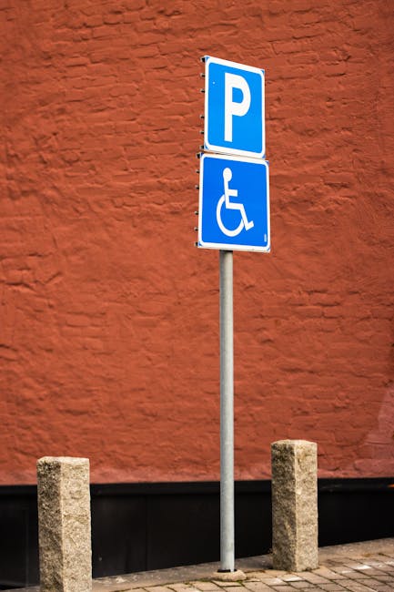 A tall metal parking signpost standing on a sidewalk in front of a red brick wall, featuring two blue signs: one indicating general parking and the other displaying a wheelchair symbol for accessible parking. The signpost is flanked by two short concrete bollards positioned on the pavement, which is made of grey concrete slabs. In the background, the textured red brick wall provides a vivid contrast to the blue signs. The image captures a typical urban parking area, possibly near residential or commercial premises, suitable for facilitating home relocation or furniture transport services by Man with Van Morden Park, with visible elements indicating parking and accessibility considerations for moving logistics.