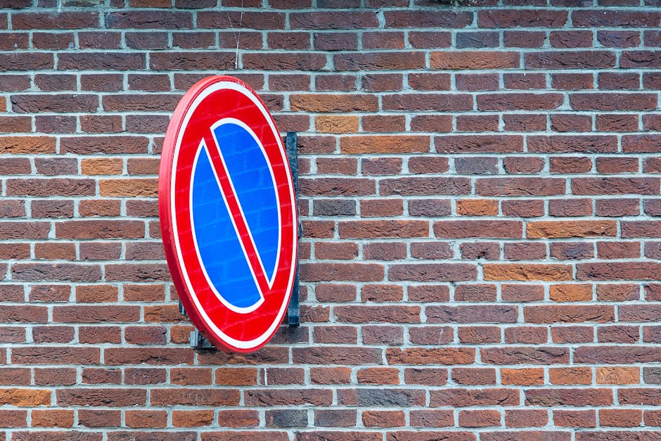A circular no parking sign mounted on a black metal bracket is attached to a red brick wall. The sign features a blue background with a red border and a prominent red diagonal line across the center, indicating parking restrictions. The brick wall behind the sign has a traditional pattern of horizontally laid bricks in shades of red, brown, and orange, with light gray mortar in between. This scene reflects a typical urban or residential exterior environment where parking regulations may impact home relocation activities. At the site, there are no vehicles, furniture, packaging materials, or moving equipment visible, but the context suggests that the sign may be relevant to managing parking suspensions during house removals or furniture transport to avoid fines and facilitate smooth moving logistics, as outlined by Man with Van Morden Park for services such as removals and home relocation.