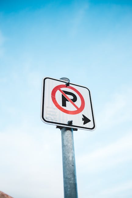 A tall metal parking restriction sign mounted on a silver pole against a clear blue sky, indicating no parking with a black letter 'P' crossed out in red and an arrow pointing right; the sign is situated near a residential area, which may relate to regulations impacting home relocations or furniture transport for Morden Park removals by Man with Van Morden Park. The image highlights the importance of understanding parking suspension rules during house moving processes to avoid fines, especially in residential streets where parking restrictions are enforced. It provides a neutral, factual visual relevant to moving services, emphasizing the need for careful planning around local parking policies when scheduling moves or home relocation logistics.