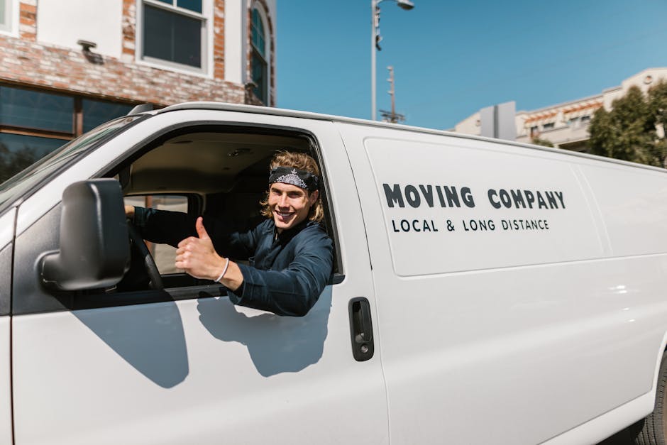 A smiling male mover with long hair wearing a black headband and dark clothing sits in the driver’s seat of a white van, giving a thumbs-up gesture. The van, branded with black lettering reading 'MOVING COMPANY LOCAL & LONG DISTANCE,' is parked outside a residential property during daytime. The background features a modern house with large windows, a brick facade, and a blue sky, indicating a home relocation scene. The vehicle is positioned near an entrance or driveway, and the driver is ready to assist with furniture transport and packing and moving logistics as part of a house removal service provided by Man with Van Morden Park.