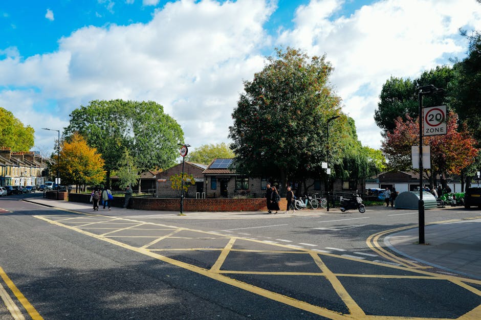 A wide view of a residential street in Morden Park showing a small community square with several pedestrians walking on the pavement. In the center, there is a brick wall surrounding a large mature tree with green and reddish leaves, indicating an autumn season. Behind the wall, low-rise buildings are visible, with some roof-mounted solar panels. The street is lined with cars parked along the edges, and bicycles are secured to bike racks near the pavement. Two black lampposts and a road sign indicating a 20 mph speed zone are visible, along with yellow road markings including a box junction and double yellow lines. The scene is well-lit with natural daylight, under a partly cloudy sky. The photo captures elements typical of a neighbourhood suitable for residential relocations, with potential involvement of [COMPANY_NAME] for home moving and furniture transport services, particularly during the process of packing, loading, and transport in house removals.