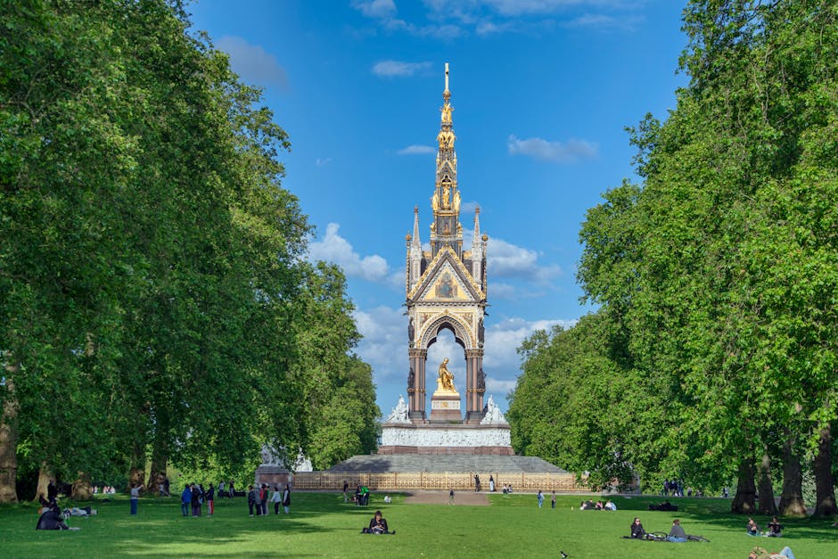 A wide view of a monument situated in a park with lush green trees on either side and a clear blue sky above. The monument features a tall, ornate spire with intricate details, a golden statue at its base, and several smaller decorative elements. People are sitting on the grass and walking around the area, with some near the base of the monument and others in the open green space. The setting suggests a peaceful environment suitable for outdoor relaxation and sightseeing, while the scene indirectly relates to home relocation or moving activities by emphasizing the importance of organized space and outdoor surroundings, which are relevant for professional removals and furniture transport services provided by Man with Van Morden Park.