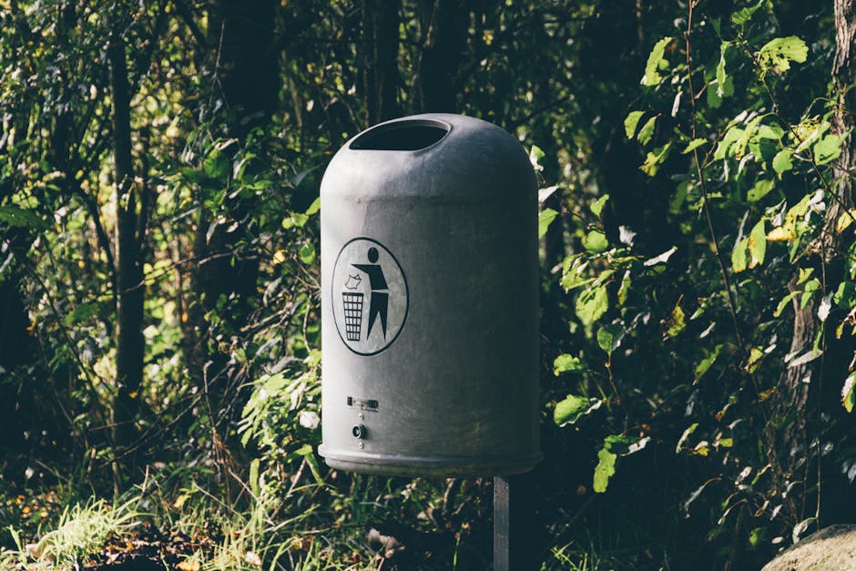 A grey cylindrical outdoor trash bin with a rounded lid and an open slot for waste collection, situated on a paved or dirt surface next to green foliage and bushes. The bin features a black and white pictogram depicting a person discarding rubbish into the bin, indicating its purpose for waste disposal, often used in parks or outdoor areas near homes during relocation or clearing activities. The scene appears to be well-lit by natural sunlight, highlighting the bin's surface and surrounding plant life, which includes leafy branches and grass. The background consists of dense greenery, and the bin is positioned in an outdoor setting possibly near an entrance or pathway. This image relates to waste disposal rules in Morden Park during home clearance or moving processes, as managed or advised by Man with Van Morden Park, a professional removals service provider.